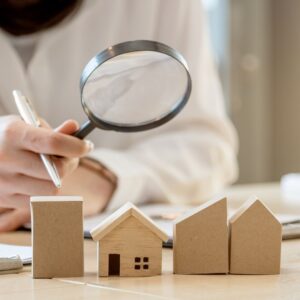 Person holding a magnifying glass over four small model homes and buildings. 